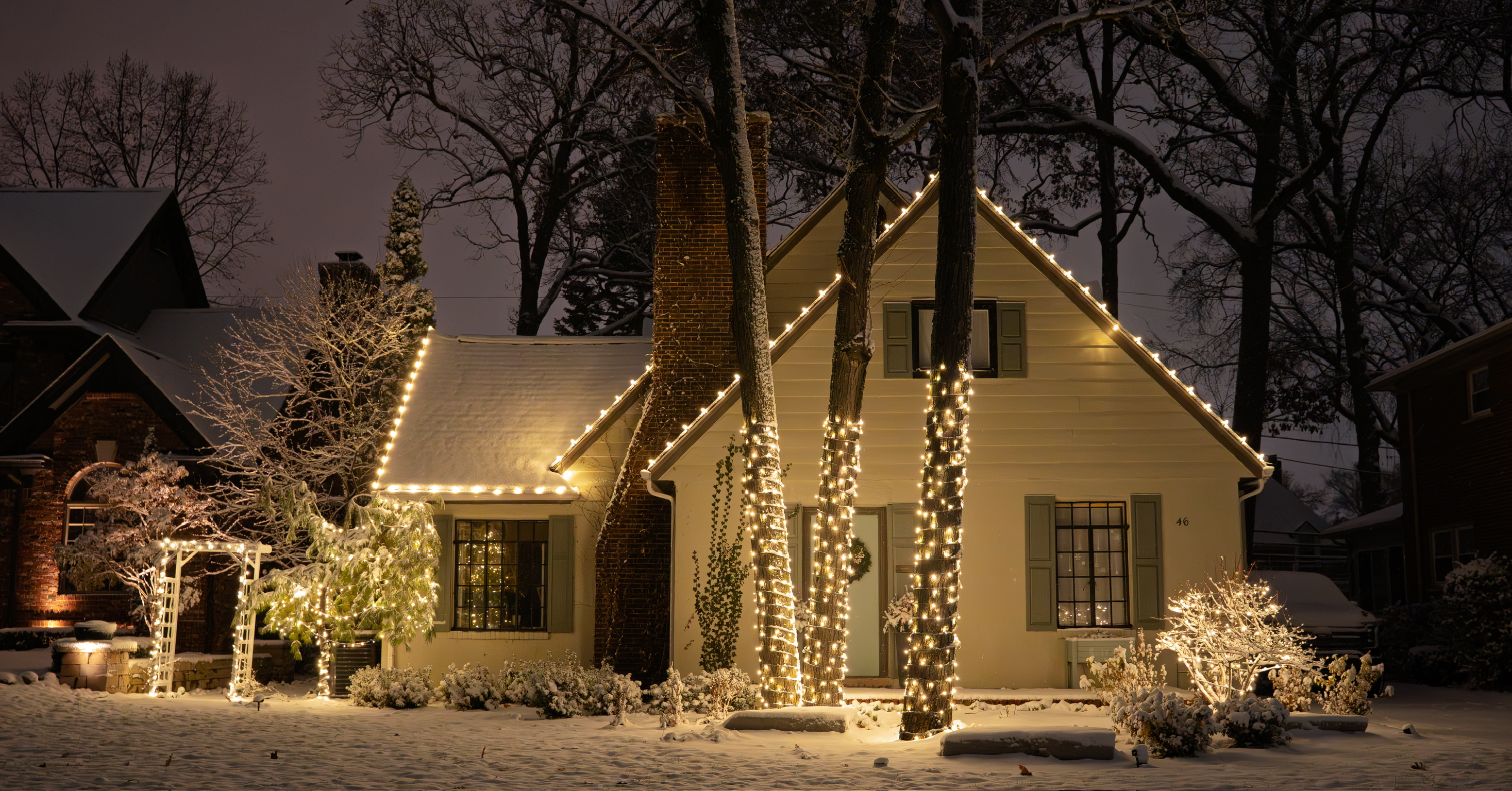 A snow-covered home glowing with warm white holiday lights along the roofline and around tall trees
