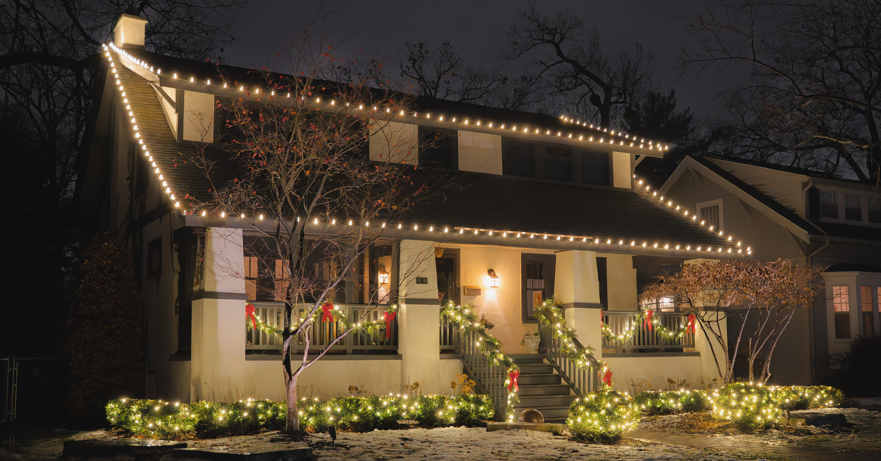 Craftsman home with warm roofline lights, glowing shrubs, and garlands with red bows