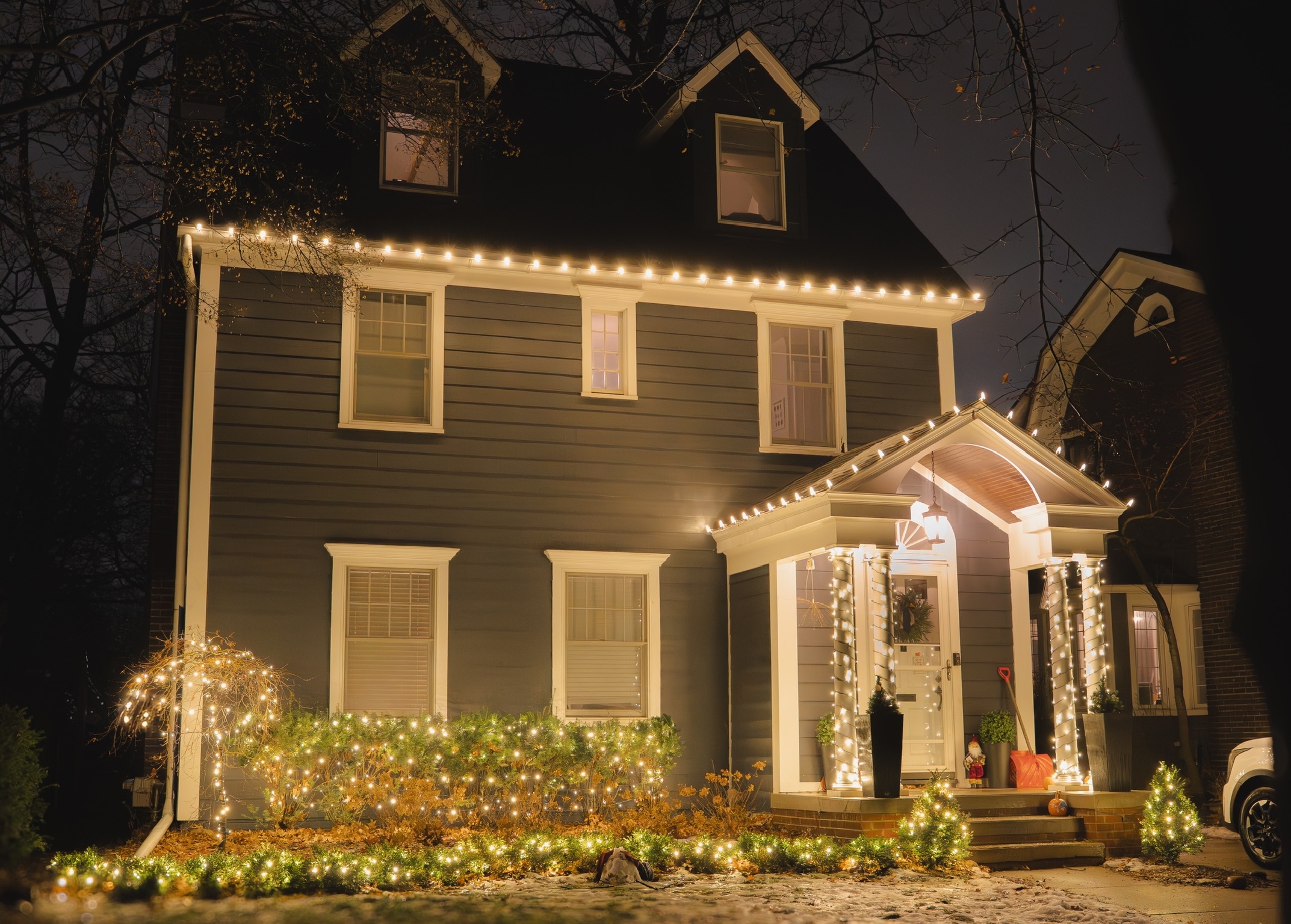 Home with warm white roofline lighting, wrapped porch columns, and glowing garden shrubs