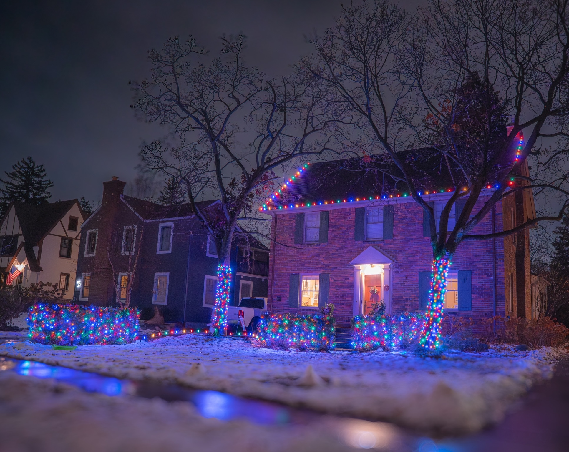 Brick home with multicolored lights along the roofline, trees, and shrubs in the snow