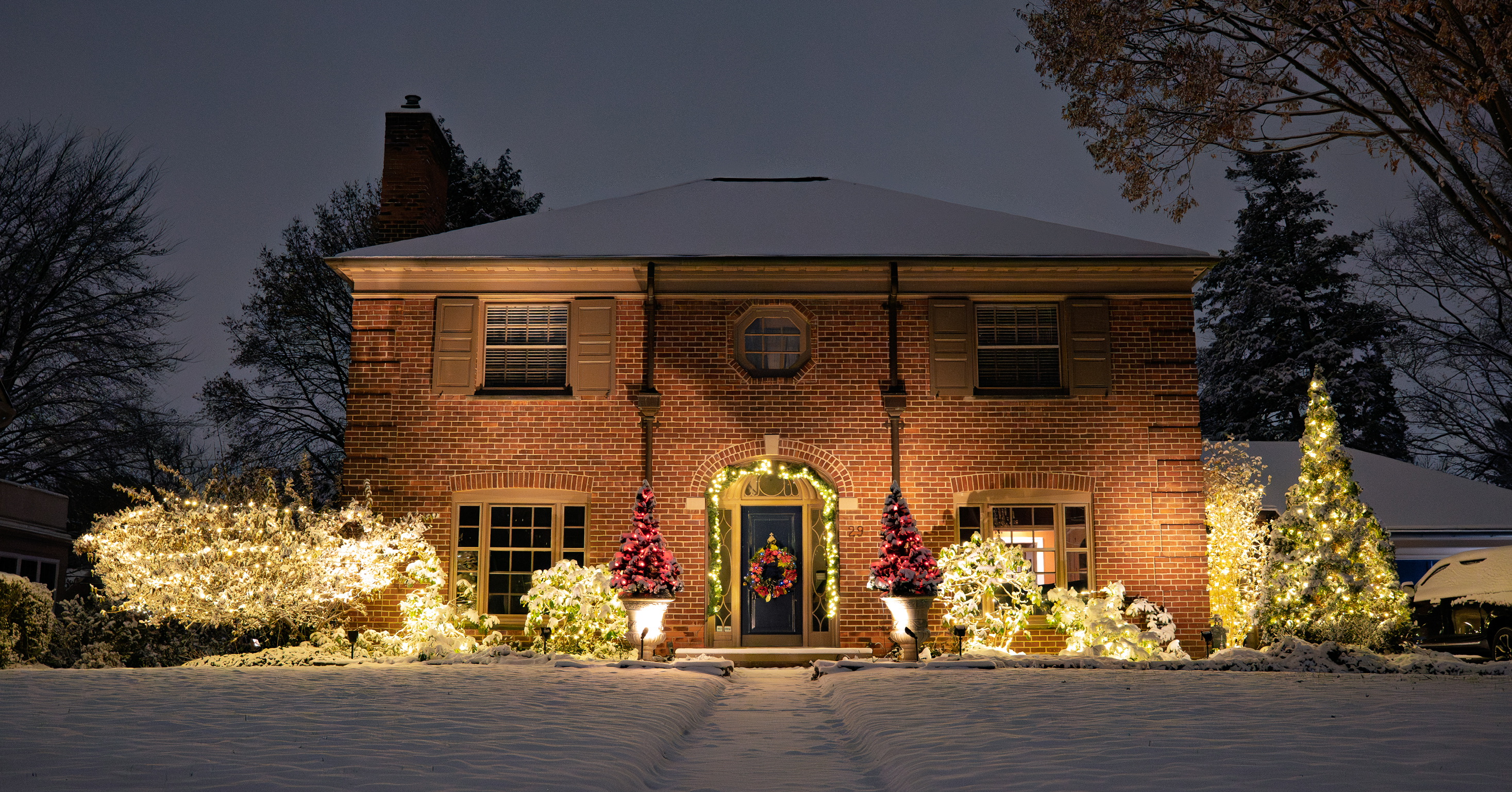 Snow-covered brick home with an illuminated entryway, shrubs, and evergreen trees