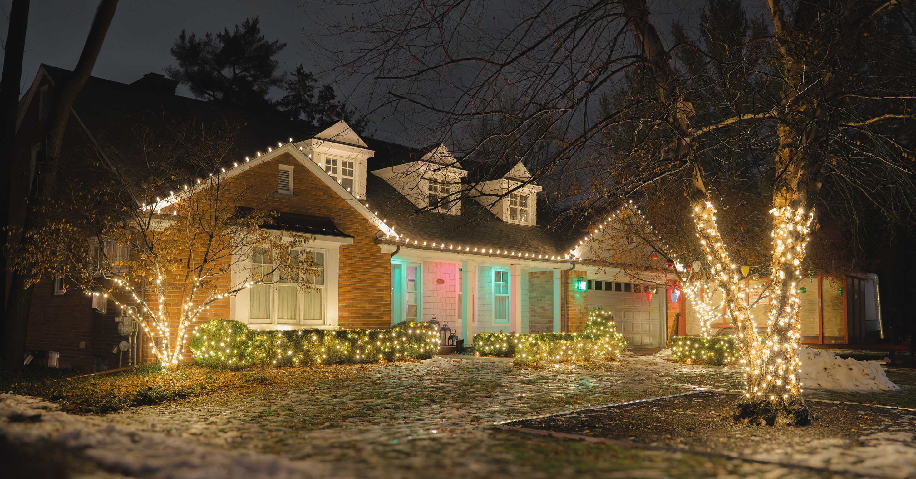 Home with warm white roofline lights, illuminated shrubs, and wrapped trees